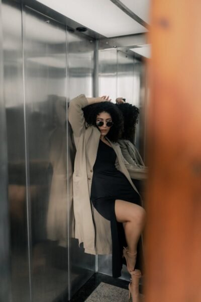Fashionable woman in a trench coat and sunglasses posing in an elevator, exuding confidence.
