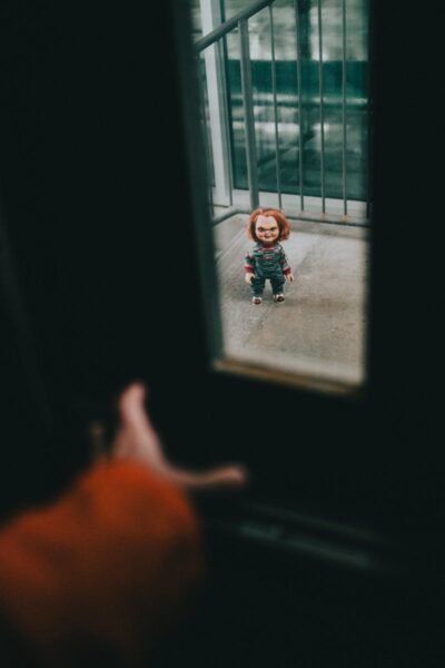 A doll with red hair stands outside a glass door, creating an eerie atmosphere.