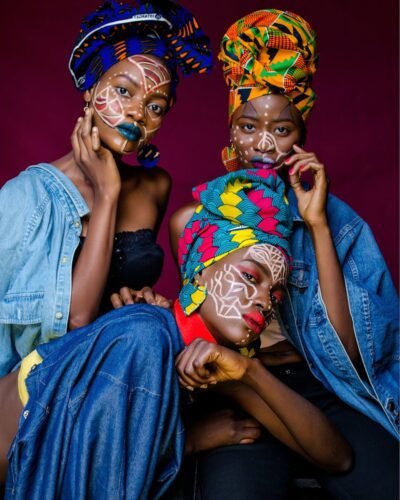 Three women in colorful African outfits with face paint and headdresses celebrating culture.