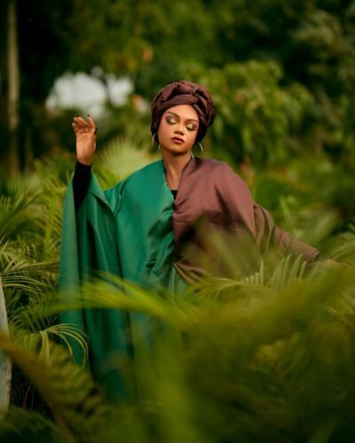 Woman in vibrant outfit, surrounded by lush green foliage, eyes closed and hand raised.
