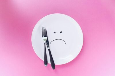A plate with a sad face drawing and cutlery on a vibrant pink background symbolizes sadness.
