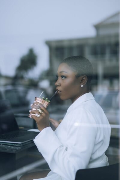 Stylish woman in white sipping a drink while working on a laptop indoors.