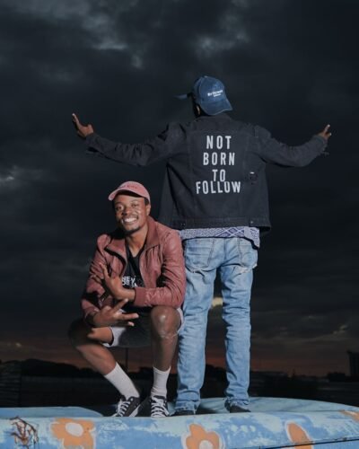 Two stylish young men pose with a bold statement jacket against a dramatic night sky.