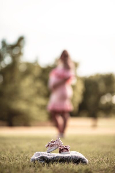 Blurry pregnant woman holding baby shoes in a sunlit park setting.